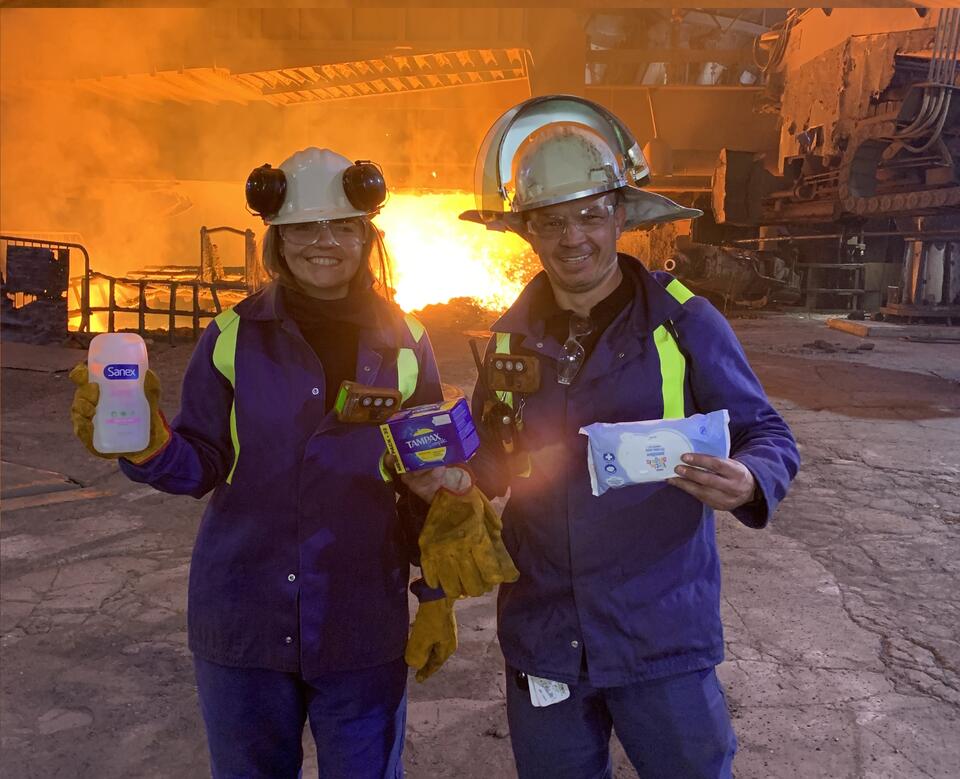 Two steel workers in the forefront, holding sanitary products, with the steel making process in the background Two steel workers in the forefront, holding sanitary products, with the steel making process in the background