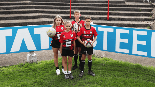 Two young girls (left) and two young boys (right) stand facing the camera on a grassy pitch dressed in red and black Tata Steel branded sports kit. The tallest girl (behind) holds a netball and the two boys on the right hold rugby balls. Light marine Tata Steel branding can be seen in the background. Two young girls (left) and two young boys (right) stand facing the camera on a grassy pitch dressed in red and black Tata Steel branded sports kit. The tallest girl (behind) holds a netball and the two boys on the right hold rugby balls. Light marine Tata Steel branding can be seen in the background.