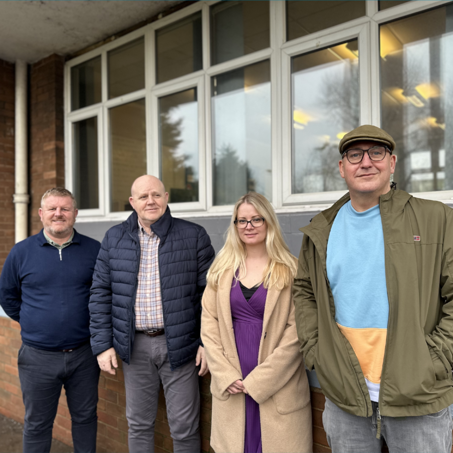 Three men and a woman stand and smile outside a brick building Three men and a woman stand and smile outside a brick building