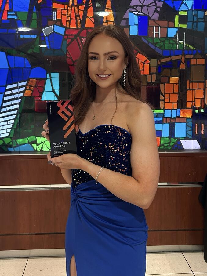 Georgia Cox wears blue gown and smiles, holding Wales STEM Award