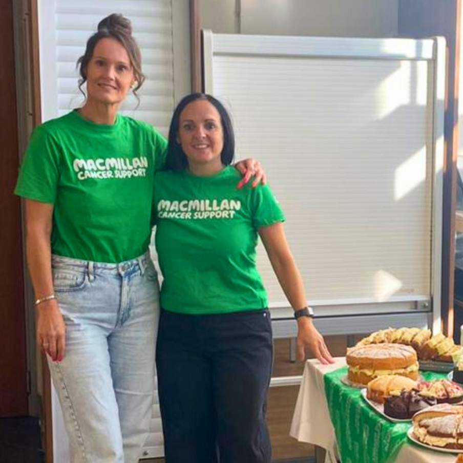 Two female steelworkers dressed in green Macmillan t shirts smile standing next to cake table